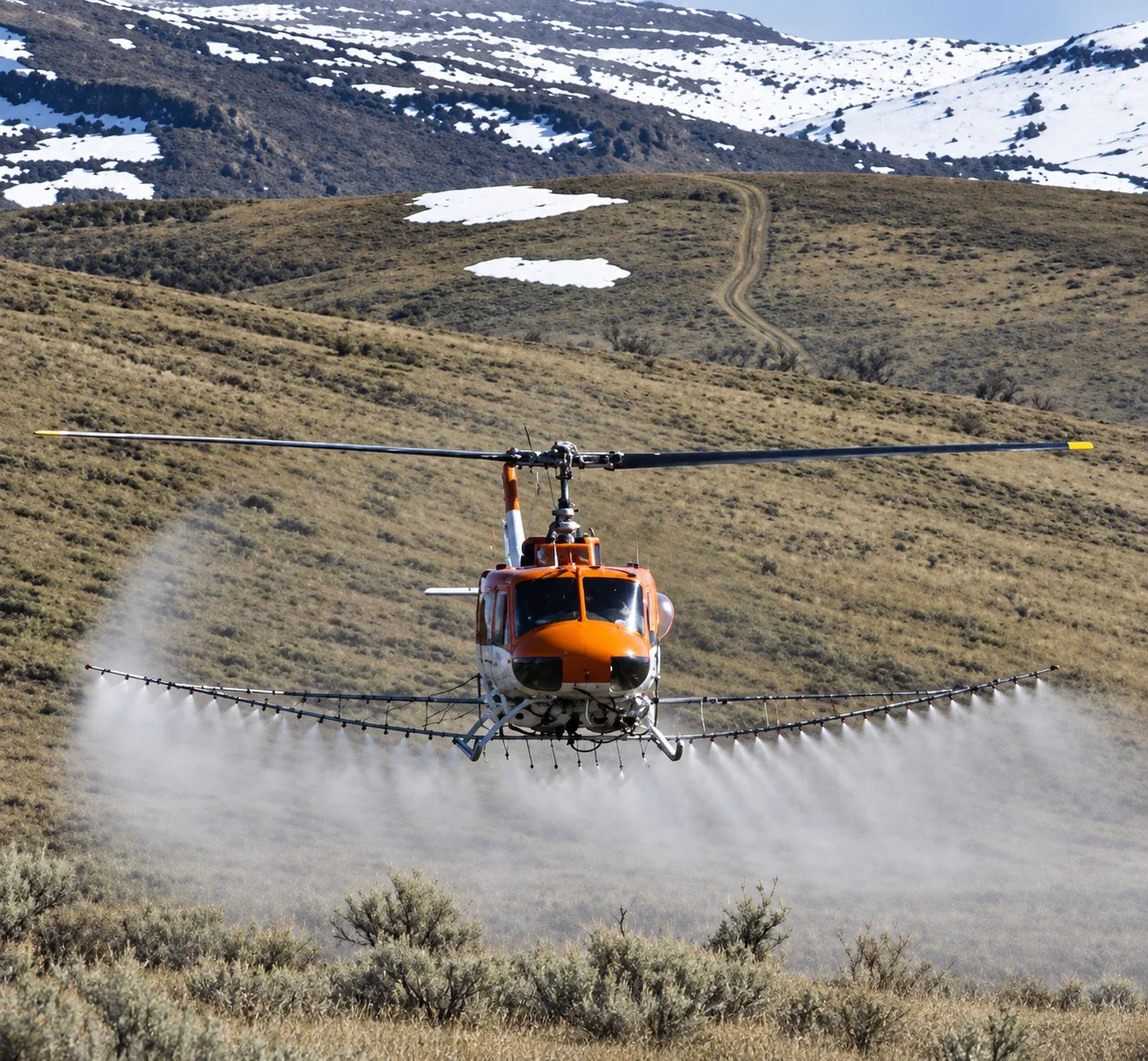 Helicopter spraying sagebrush rangeland below snow-covered hills.